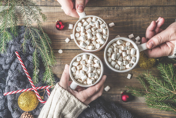Winter partij concept met warme chocolademelk, drie handen met cacao cups met marshmallow op houten tafel met kerstversiering, top indeling kopie space bekijken - Foto, afbeelding