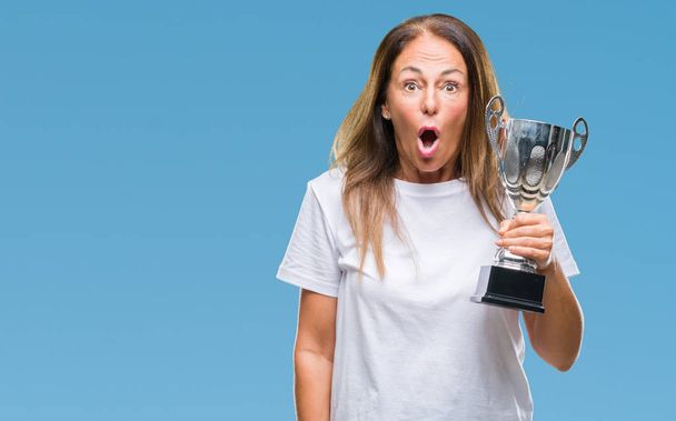 Mulher vencedora hispânica de meia-idade celebrando prêmio segurando troféu sobre fundo isolado assustado em choque com um rosto surpresa, com medo e animado com expressão de medo
 - Foto, Imagem