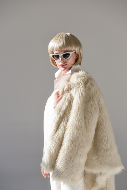 portrait of attractive blonde woman in sunglasses and fashionable winter outfit with faux fur coat looking at camera on white - Zdjęcie, obraz