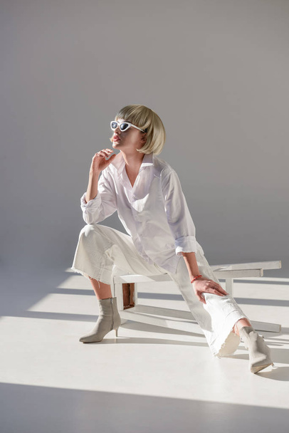 attractive blonde woman in sunglasses and fashionable white outfit sitting on chair and looking away on white - Foto, afbeelding