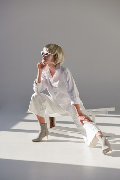 attractive blonde woman in sunglasses and stylish white outfit sitting on chair and looking away on white - Φωτογραφία, εικόνα