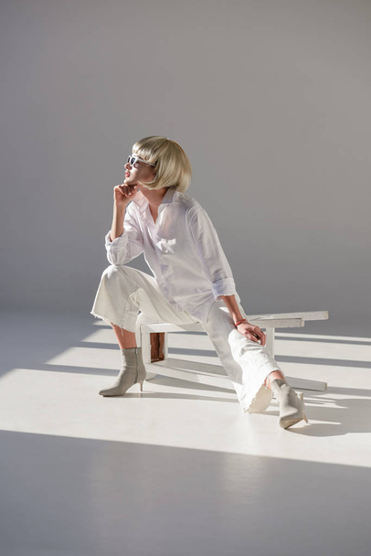 attractive blonde woman in sunglasses and trendy white outfit sitting on chair and looking away on white - Photo, Image