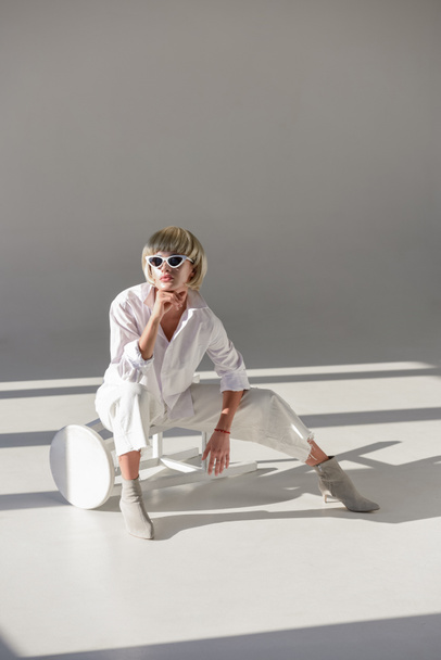 attractive blonde woman in sunglasses and fashionable white outfit sitting on chair and resting chin on hand on white - Foto, immagini