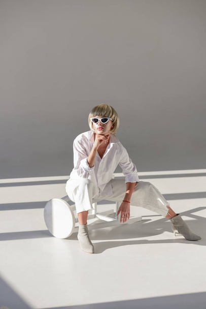 beautiful girl in sunglasses and fashionable white outfit sitting on chair and resting chin on hand on white - Foto, afbeelding