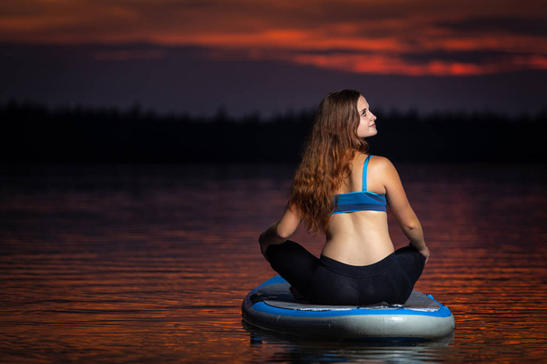 Красива струнка дівчина з довгим коричневим волоссям займається йогою на дошці в темному, барвистому заході сонця на мальовничому озері
 - Фото, зображення