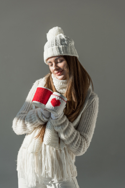 cheerful attractive woman in fashionable winter sweater and scarf holding two cups isolated on grey - Photo, Image
