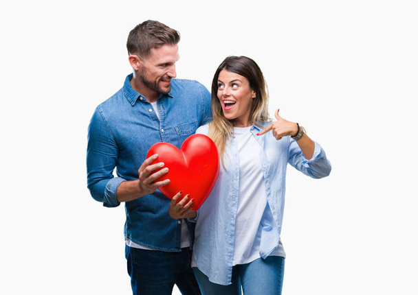 Молодая влюбленная пара держит красное сердце на изолированном фоне с удивленным лицом, указывающим пальцем на себя
 - Фото, изображение