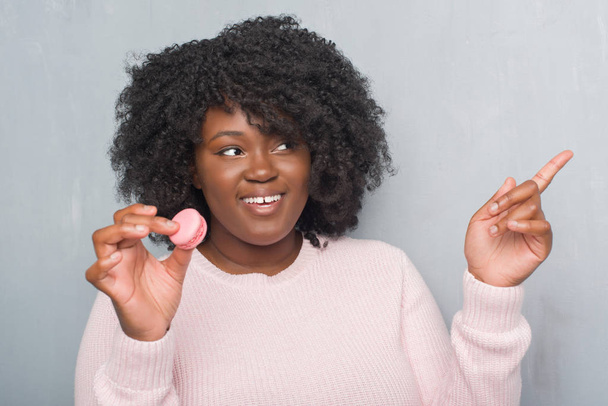 Fiatal afrikai-amerikai nő evés rózsaszín macaron nagyon boldog, mutat, a kéz és az ujjak oldalán szürke grunge falon - Fotó, kép