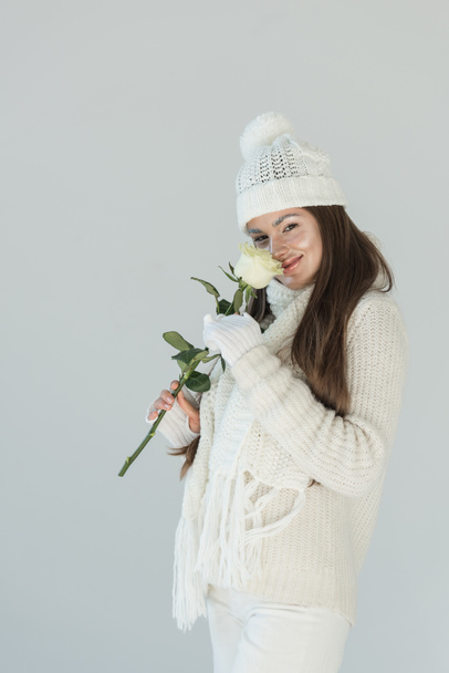 ελκυστική γυναίκα μόδας χειμώνα πουλόβερ και κασκόλ sniffing λευκό τριαντάφυλλο και βλέπουν τα φωτογραφικών μηχανών που απομονώνονται σε λευκό - Φωτογραφία, εικόνα