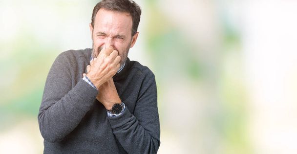 Knappe middenleeftijd senior man dragen een trui over geïsoleerde achtergrond ruiken iets stinkende zijn en walgelijk, ondraaglijke geur, adem met vingers op de neus. Slechte geuren concept. - Foto, afbeelding