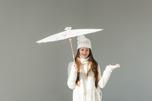 attractive woman in fashionable winter sweater and scarf standing under paper umbrella isolated on grey - Zdjęcie, obraz