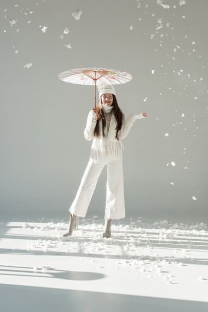 happy woman in fashionable winter sweater and scarf standing under japanese umbrella, snow falling on white - Foto, Imagem