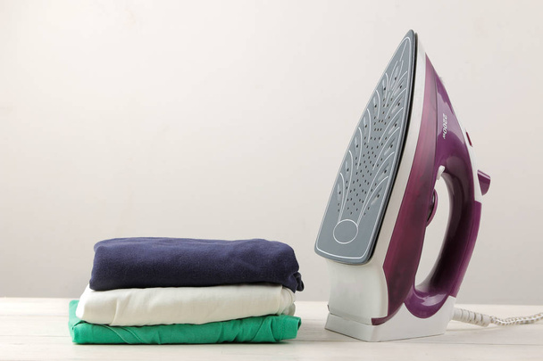 Fliederbügeleisen und ein Stapel Kleidung auf hellem Hintergrund. Wäsche bügeln. Elektrische Haushaltsgeräte. - Foto, Bild