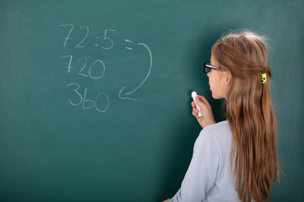 Rear View Of A Girl Wearing Eyeglasses Solving Mathematics On Chalkboard - Zdjęcie, obraz