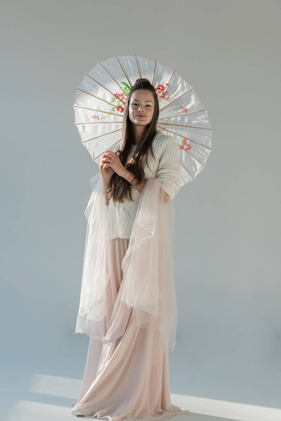 attractive woman in fashionable winter outfit standing with japanese umbrella and looking at camera on white - Fotó, kép