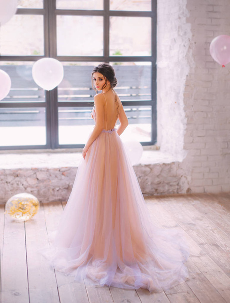A graceful young lady with dark hair in a gorgeous tender long peach and purple dress with a train and open back, turns to the camera in a bright and spacious room filled with light color balls. - Foto, Imagem