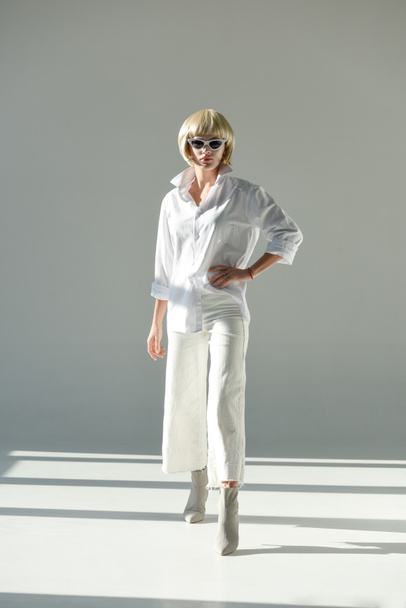 attractive blonde woman in sunglasses and fashionable white outfit with hand akimbo looking at camera on white - Фото, зображення