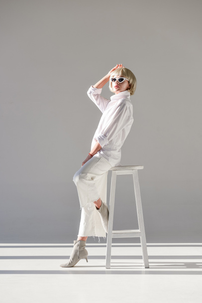 attractive blonde woman in sunglasses and fashionable white outfit sitting and posing on chair on white - Photo, Image
