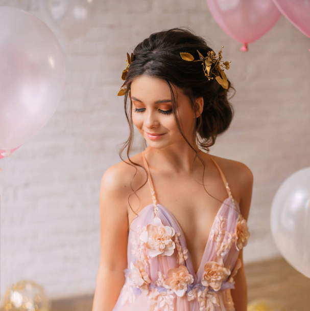 a wonderful young elf queen with dark hair and gold in a chic light pink dress with purple color, shyly closes her eyes, stands in a bright spacious room near white balloons. - Фото, изображение