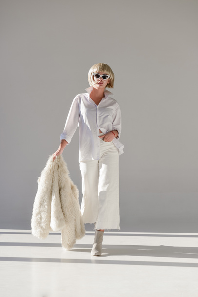 atractiva mujer rubia en gafas de sol y ropa de invierno de moda caminando con abrigo de piel sintética en blanco
 - Foto, Imagen