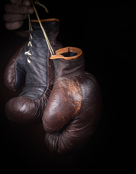 рука держит пару коричневых очень старых боксерских перчаток на нитке, черный фон
 - Фото, изображение