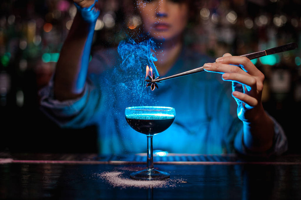 Chica camarera añadiendo a un cóctel marrón un badian flameado con pinzas en la luz azul
 - Foto, imagen