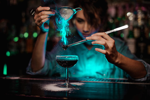 Female bartender pouring on the brown cocktail and on a flamed badian on tweezers a powdered sugar in the green light - Zdjęcie, obraz