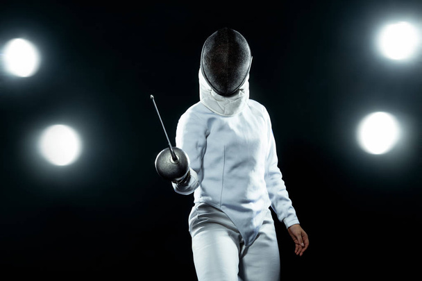 Young fencer athlete wearing fencing costume holding the sword and mask. Isolated on black background with lights - Fotoğraf, Görsel