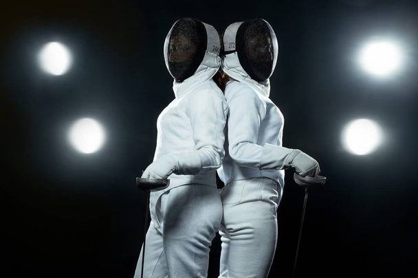 Young fencer athlete wearing mask and white fencing costume. holding the sword on black background with lights. - Photo, Image