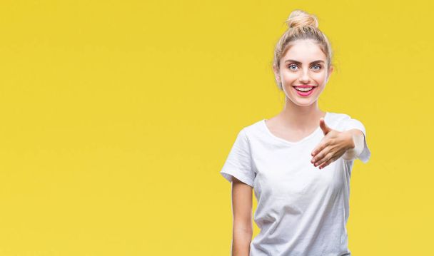 Jeune belle femme blonde portant un t-shirt blanc sur fond isolé souriant offrant une poignée de main pour saluer et accueillir. Une entreprise prospère
. - Photo, image