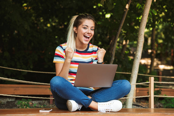 Portrait of an excited young teenage girl sitting on a bench at the park, using laptop computer on her lap, celebrating - 写真・画像