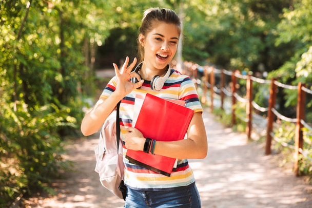 Happy young teenage girl carrying backpack and textbooks walking at the park - Fotoğraf, Görsel