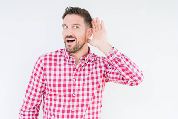 Jonge knappe man shirt dragen over geïsoleerde achtergrond glimlachend met hand over oor luisteren een hoorzitting gerucht of roddel. Doofheid concept. - Foto, afbeelding