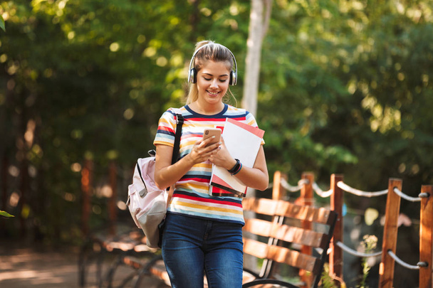 Happy young teenage girl carrying backpack and textbooks walking at the park, listening to music with headphones, using mobile phone - Foto, imagen
