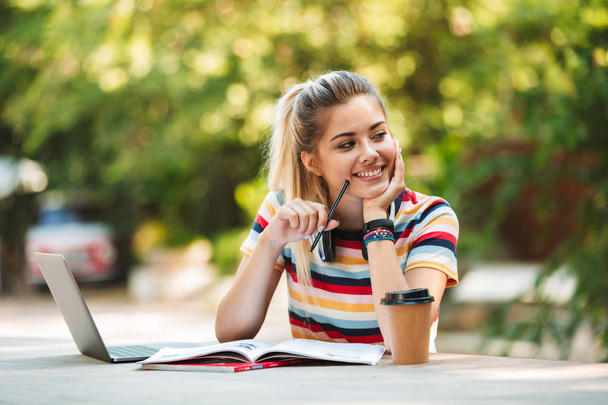 Image of a happy young cute girl student sitting in park using laptop computer writing notes. - Foto, Imagem