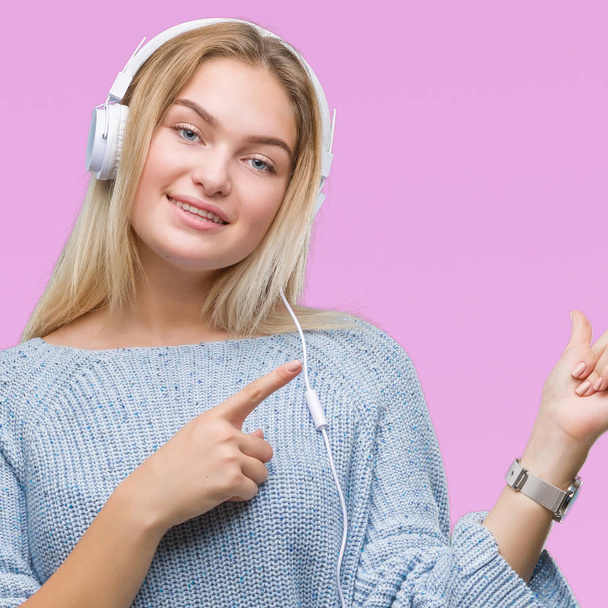Молодая белая женщина слушает музыку в наушниках на изолированном фоне улыбается и смотрит в камеру, указывая двумя руками и пальцами в сторону
. - Фото, изображение