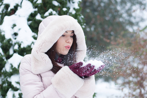 Portrait of young beautiful emotional woman in hooded down coat and gloves on snow covered garden background. Winter snowy landscape - Photo, Image