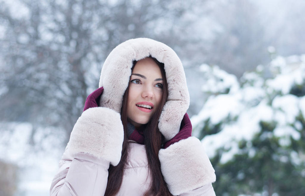 Портрет молодой красивой эмоциональной женщины в капюшоне и перчатках на заснеженном фоне сада. Зимний снежный пейзаж
 - Фото, изображение