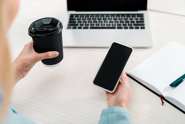 Teilbild einer Geschäftsfrau mit Coffee to go und Smartphone mit leerem Bildschirm am Arbeitsplatz - Foto, Bild