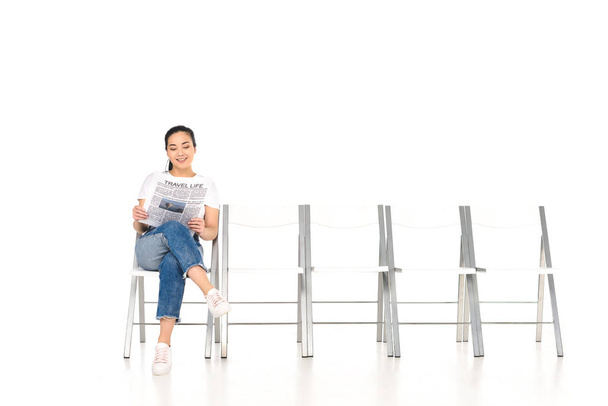 lächelndes Mädchen sitzt auf Stuhl mit überkreuzten Beinen und liest Zeitung isoliert auf weißem Papier - Foto, Bild