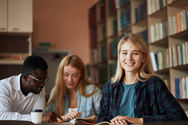 Smiling attractive blonde girl makes some notes while reading book in college library, looking at camera, sitting at desk with the other students on background - Photo, image