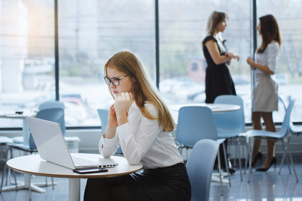 Team von Büroangestellten. Ein langhaariges Mädchen mit Brille und weißer Bluse sitzt an einem Tisch mit Laptop, im Hintergrund stehen zwei Geschäftsfrauen und unterhalten sich. Teamwork. Gleichberechtigung der Geschlechter. - Foto, Bild