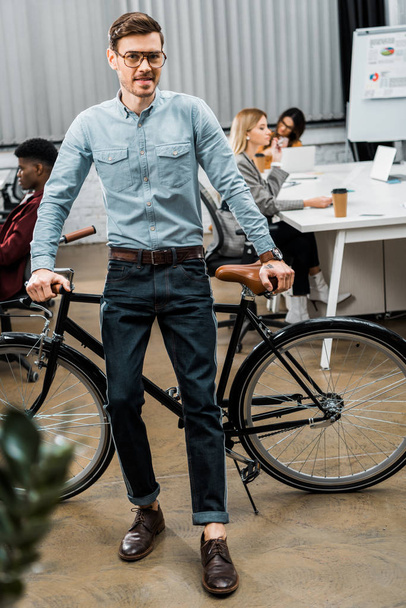 selective focus of young businessman leaning on bicycle in office with multiracial colleagues behind - Фото, зображення