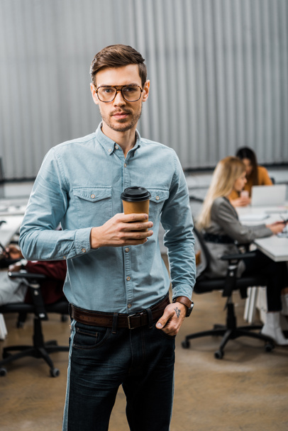 selective focus of young businessman with coffee to go in office with multiracial colleagues behind - Foto, Imagen