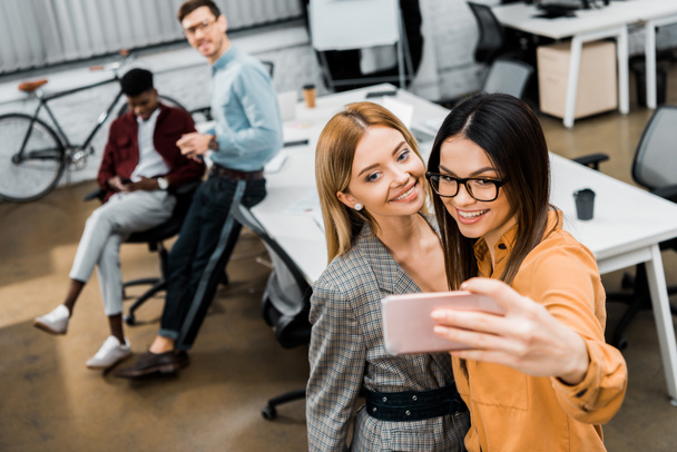 sonrientes empresarias tomando selfie en el teléfono inteligente en la oficina con colegas multirraciales detrás
 - Foto, Imagen