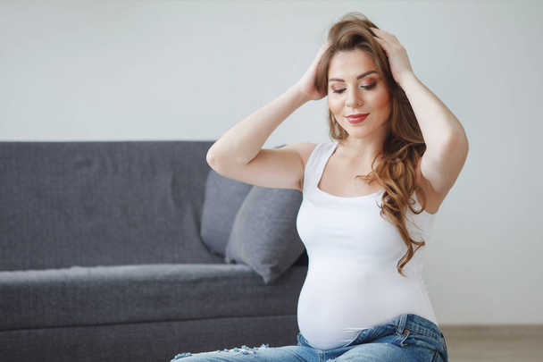 Piękna kobieta w ciąży z długimi włosami w białą kamizelkę siedzi na podłodze w pobliżu szary kanapie. Koncepcja macierzyństwo szczęśliwego, zdrowego stylu życia. - Zdjęcie, obraz