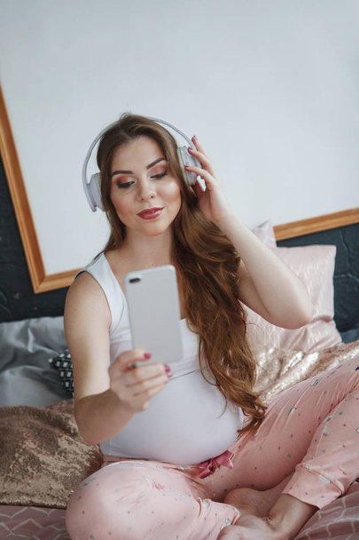 Mooie zwangere vrouw met lange haren in een wit T-shirt en roze pyjama zittend op het bed, luisteren naar muziek met een koptelefoon en kijken naar een smartphone. Concept van gelukkig moederschap, gezonde levensstijl. - Foto, afbeelding