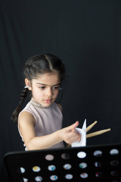 5-vuotias tyttö brittiläinen intialainen alkuperä harjoitellaan ja soittaa rumpuja, lukeminen musiikkia merkinnästä - on tumma tausta kopioi tilaa
.  - Valokuva, kuva