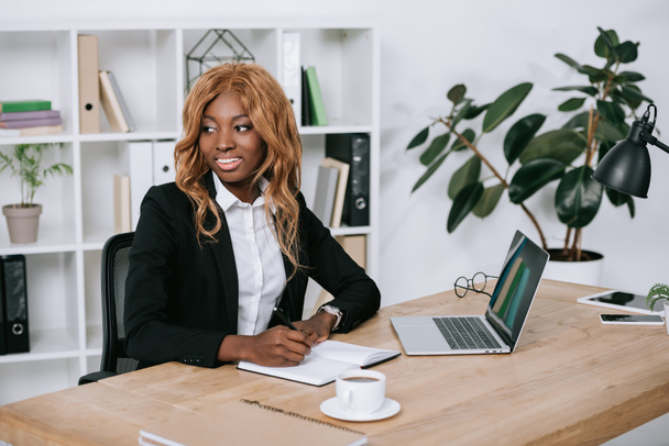 enfoque selectivo de la feliz y reflexiva mujer de negocios afroamericana escribiendo en cuaderno
  - Foto, imagen