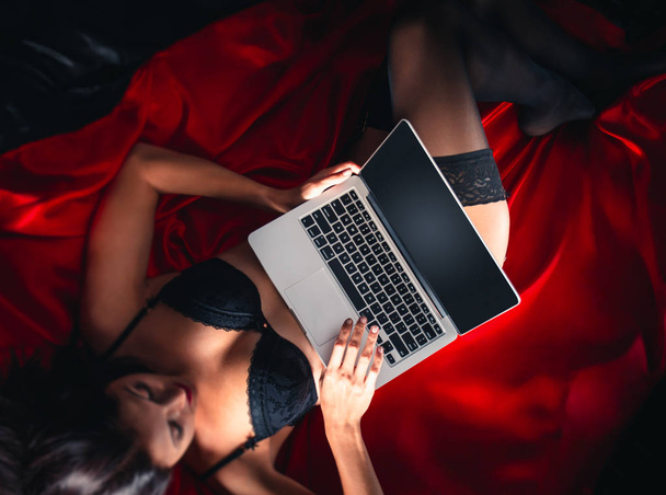 Nahaufnahme von sexy jungen brünetten Frau in schwarzen sinnlichen Dessous und in Strümpfen mit Laptop auf rotem Bett im Studio. - Foto, Bild